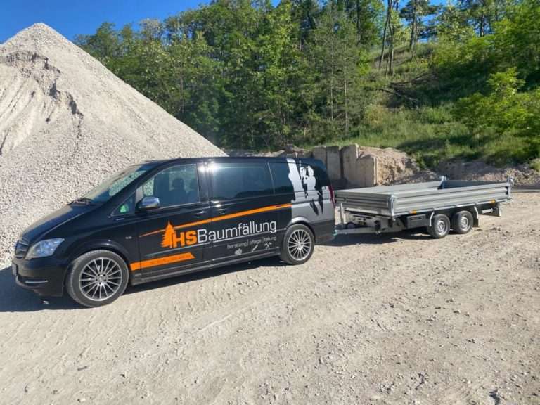 Schwarzer Transporter mit HS-Baumfällung-Logo und beladenem Kipp-Anhänger auf einem Schotterplatz
