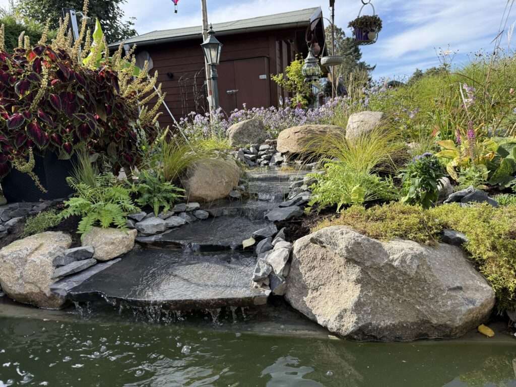 Bachlauf aus Natursteinplatten zwischen großen Findlingen, dahinter ein Gartenhaus